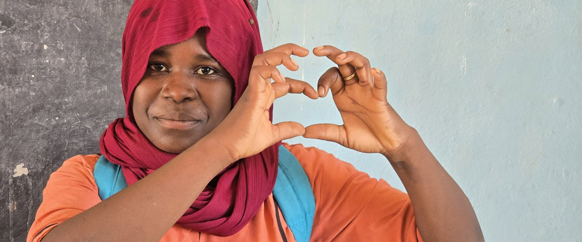 A young woman in a red hijab forms a heart shape with her hands, smiling against a light blue background.’