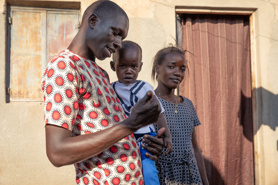 A man holds a young child while a woman stands beside them, all in front of a simple building.