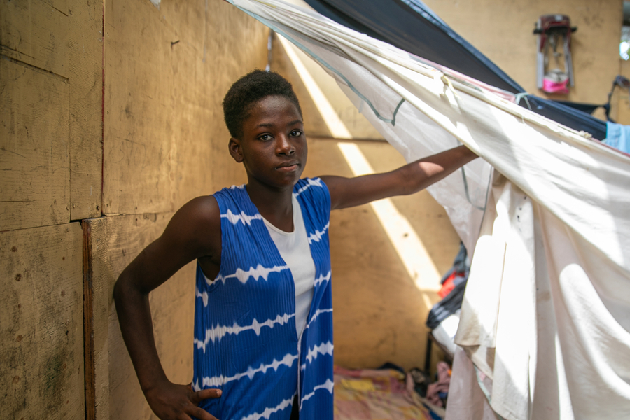 A girl wearing a long blue and white vest stands in a makeshift shelter made of plywood and white sheets.