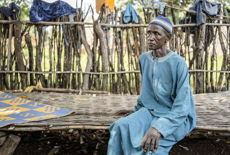 An older man wearing a blue outfit sits on a wooden bench staring to the side.