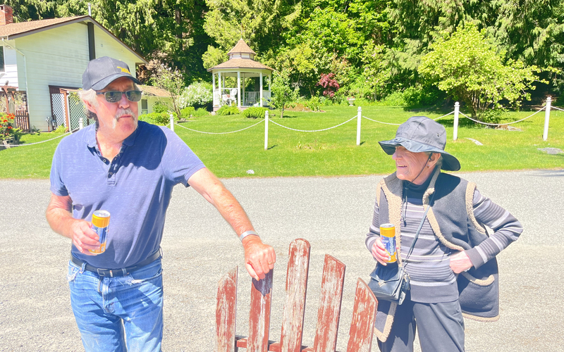A man in a blue polo shirt and a black baseball cap stands outdoors next to a red rustic wooden chair. A woman wearing black clothing and a black sun hat stands next to him. Both are holding drinks.