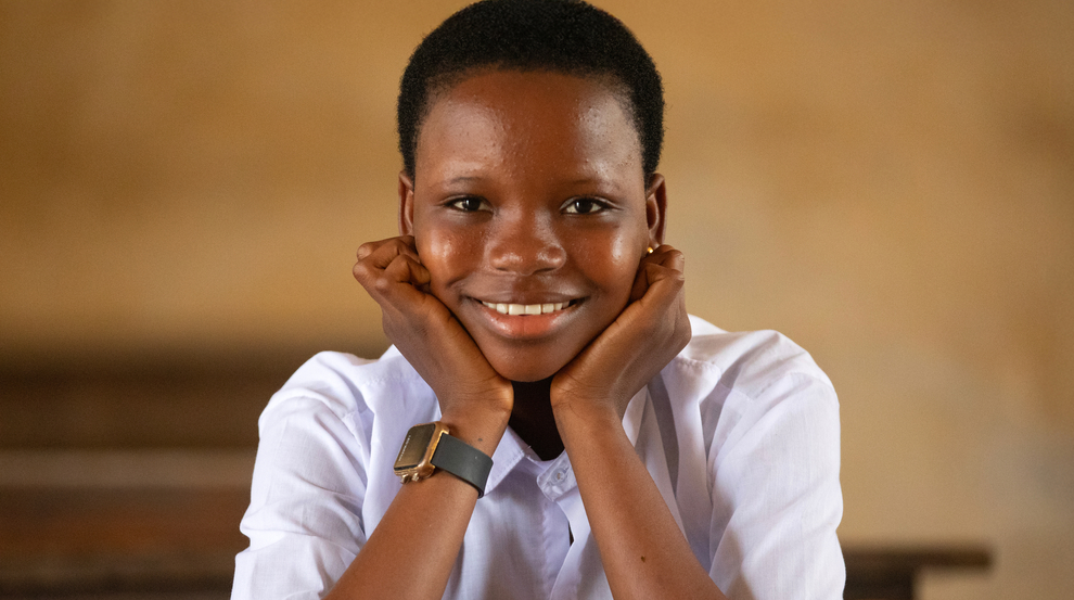 A child wearing a white shirt is sitting at a table smiling with her hands on her chin in front of a red bowl of food.