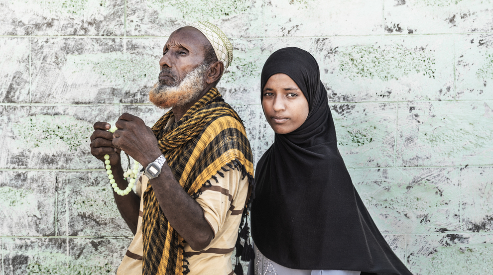 An older man with a beard wearing mostly yellow and brown stands next to a young woman wearing a black headscarf.