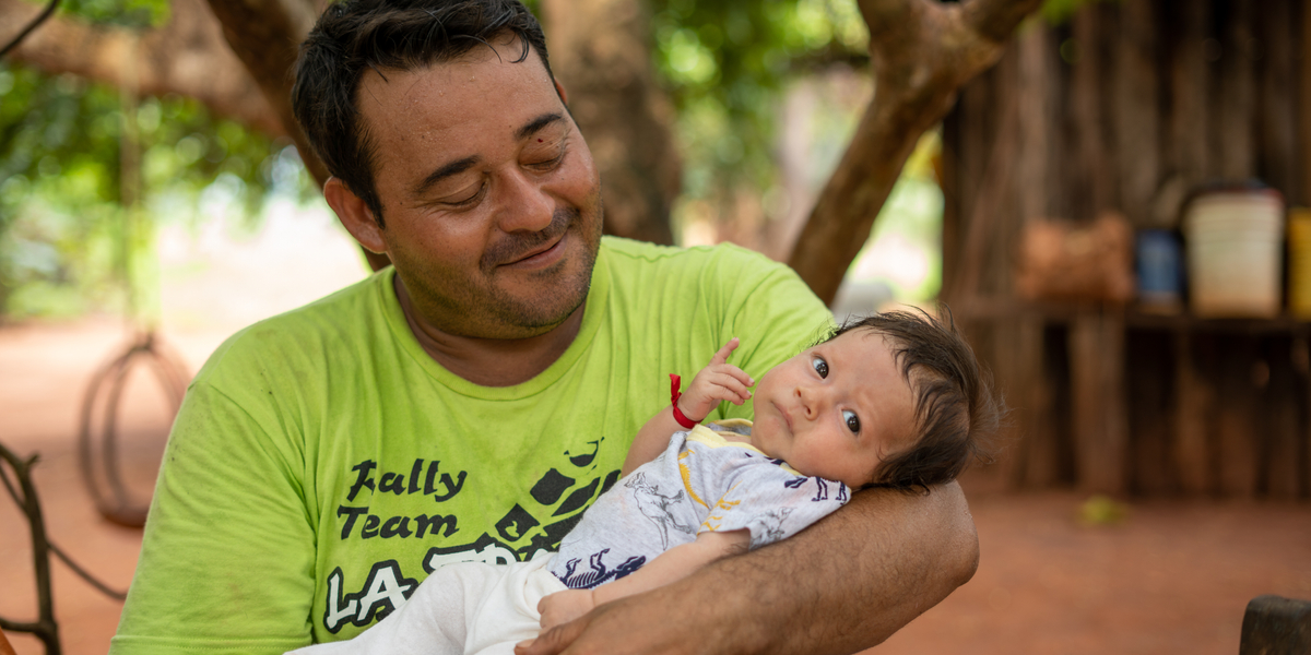 A smiling man holds a baby in his arms, surrounded by greenery and rustic wooden structures, conveying a warm, familial moment.