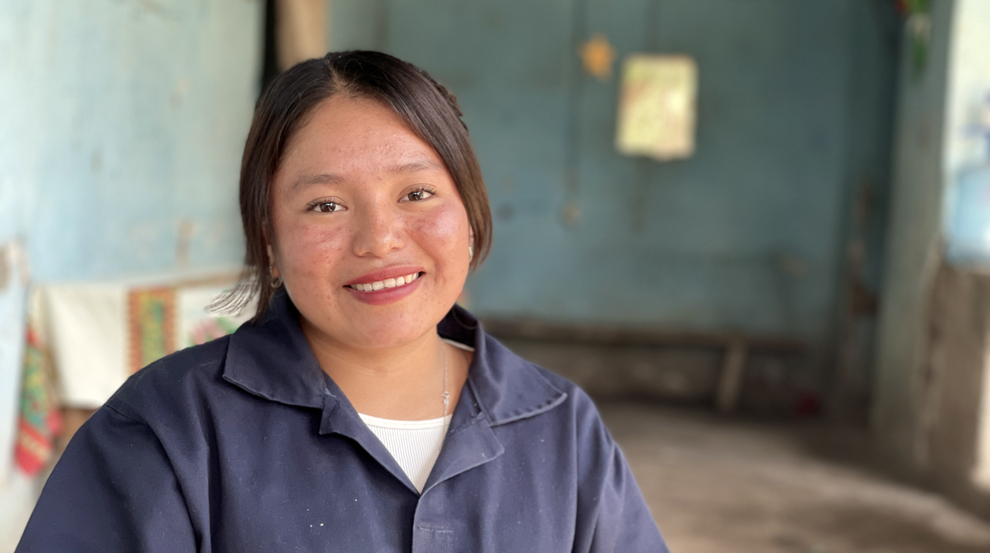 A young woman smiling in a blue jumpsuit, representing women's skills training and economic empowerment.