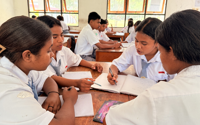 Students in school uniforms sitting together at desks, working through notes, and discussing a lesson in a classroom.
