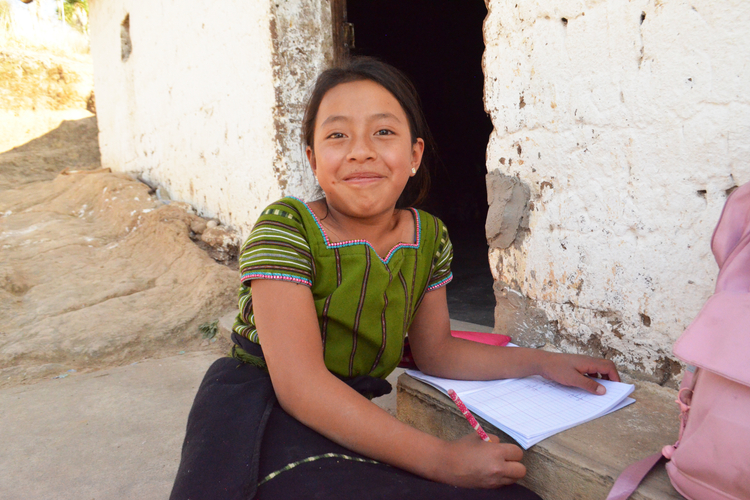 A young girl in traditional clothing sits outside a rustic building, smiling while writing in a notebook.