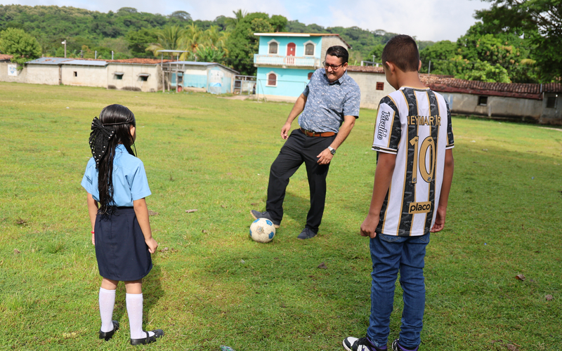 Three individuals on a grassy field playing soccer. A man kicks the ball, a young girl stands nearby, and a boy in a “NEYMAR JR 10” jersey watches. Colourful buildings and green trees are in the background.