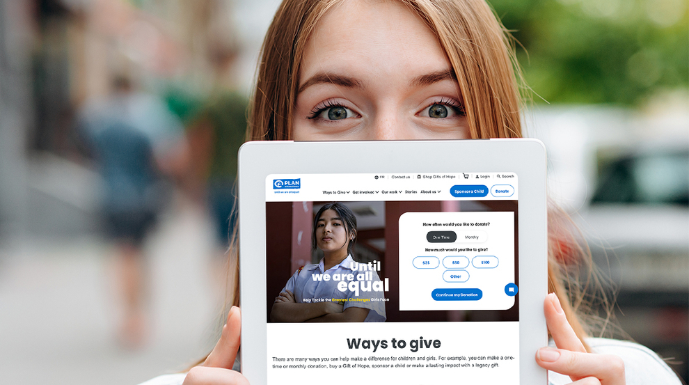 A woman with red hair and wearing a white top and light blue denim jacket holds up a tablet half covering her face with the Plan Canada website open on it.