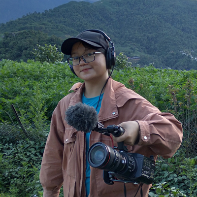 A young woman, wearing glasses, a hat, and a pink jacket, is holding a video camera and microphone.