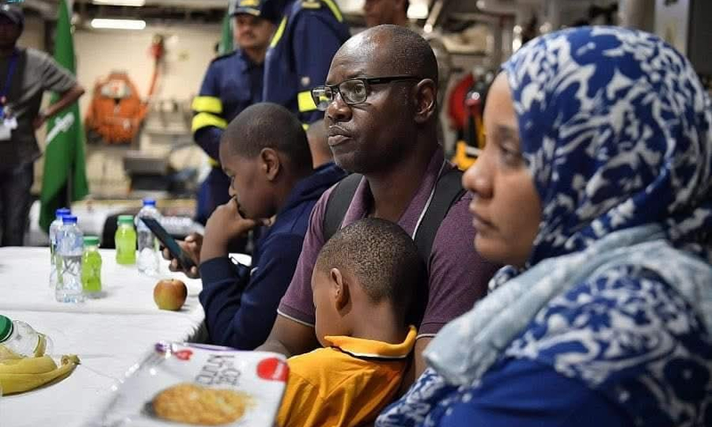 A family sits at a table with food and water bottles. The father holds his son close, while others look tired and contemplative, reflecting on the stress and hardship of their evacuation from Sudan