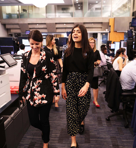 Two young women walk through a busy office workspace during a Girls Belong Here placement.