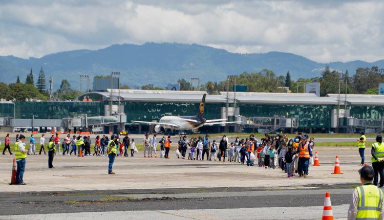 A group of adults and children standing in a line on an airport tarmac holding white plastic bags.