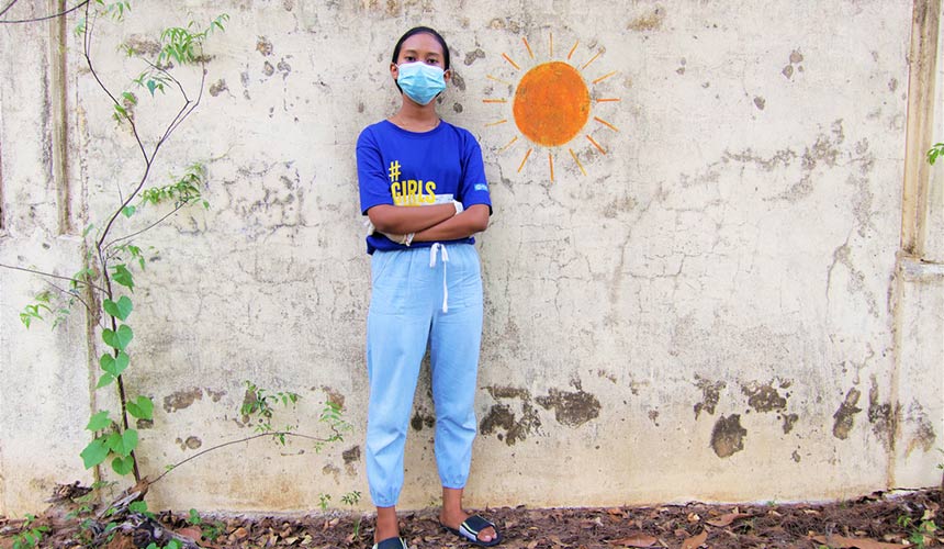 Girl in a mask standing with arms folded standing next to a Sun Symbol on a wall.