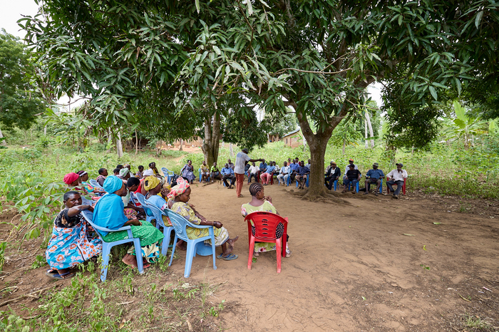 A group of people sitting on blue and red plastic chairs are sitting around a large tree listening to a man speak who is standing underneath the shade of the tree.