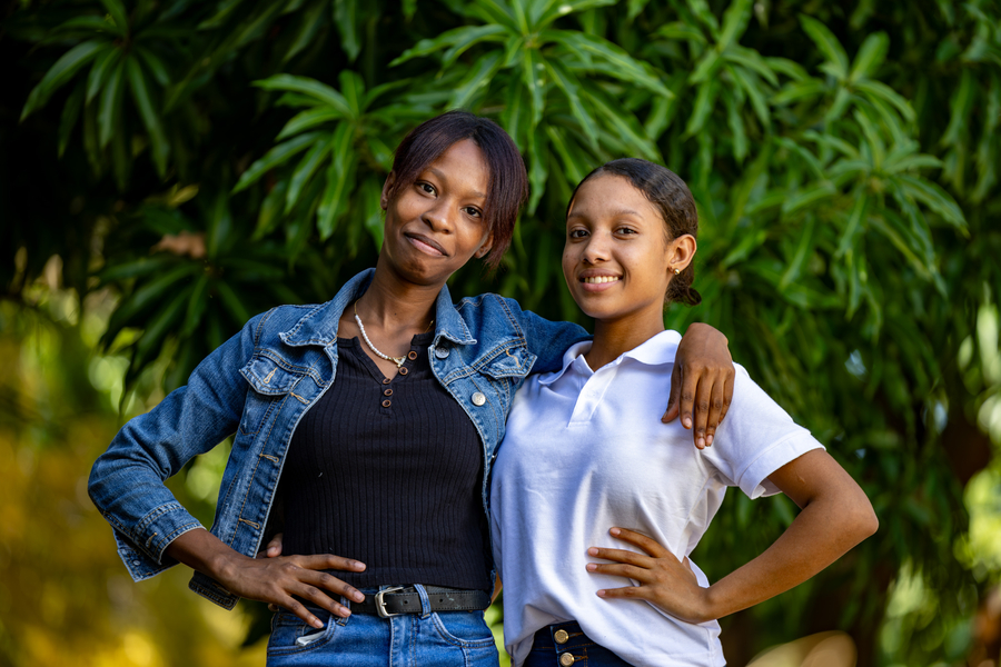Two young women stand together outdoors, smiling and posing confidently against a backdrop of lush green foliage.