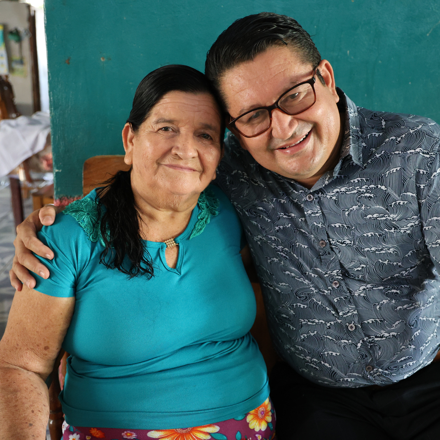 Two people sit side by side indoors on a wooden bench, one wearing a turquoise shirt with dark hair, the other wearing glasses and a patterned shirt.
