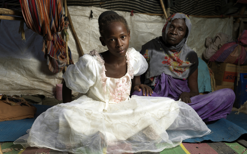 A young girl in a white dress sitting on a colorful rug with an elderly woman sitting behind her.