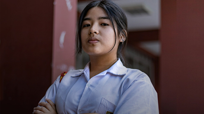 A smiling teenage girl wearing a white and pink head scarf and a white t-shirt stands on the sidewalk beside a road in rural Senegal.