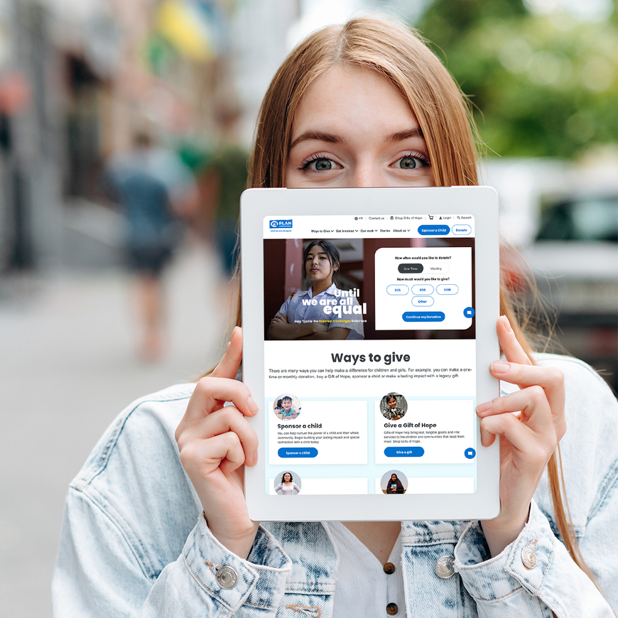 A woman with red hair and wearing a white top and light blue denim jacket holds up a tablet half covering her face with the Plan Canada website open on it.