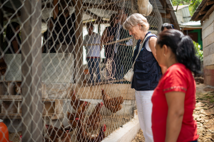 A woman with short blonde hair looks through some chicken wire at a chicken farm