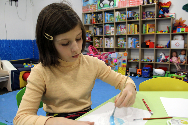 Helga, a nine-year-old girl, paints a picture at a table inside a child-friendly space.