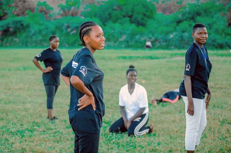 Neema, a sixteen-year-old girl, stands in a grassy field with other girls during a football game.