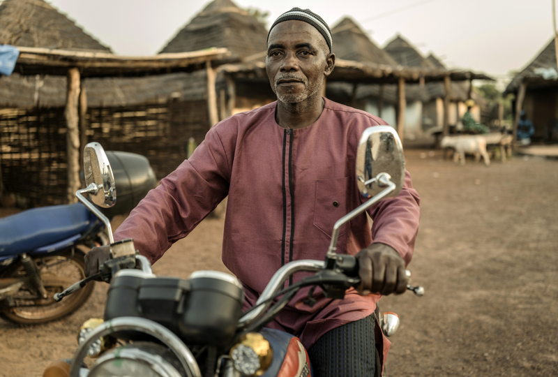 A man wearing a red jacket and black pants is riding his motorcycle. In the background there are goats and some thatched huts.