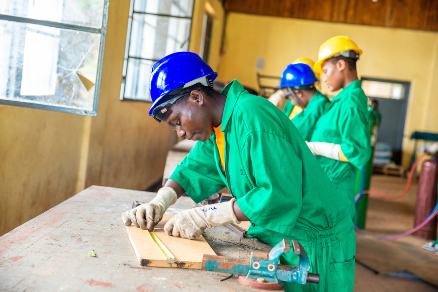 A group of students in green uniforms and safety helmets work on woodworking projects in a classroom, focused on their tasks.