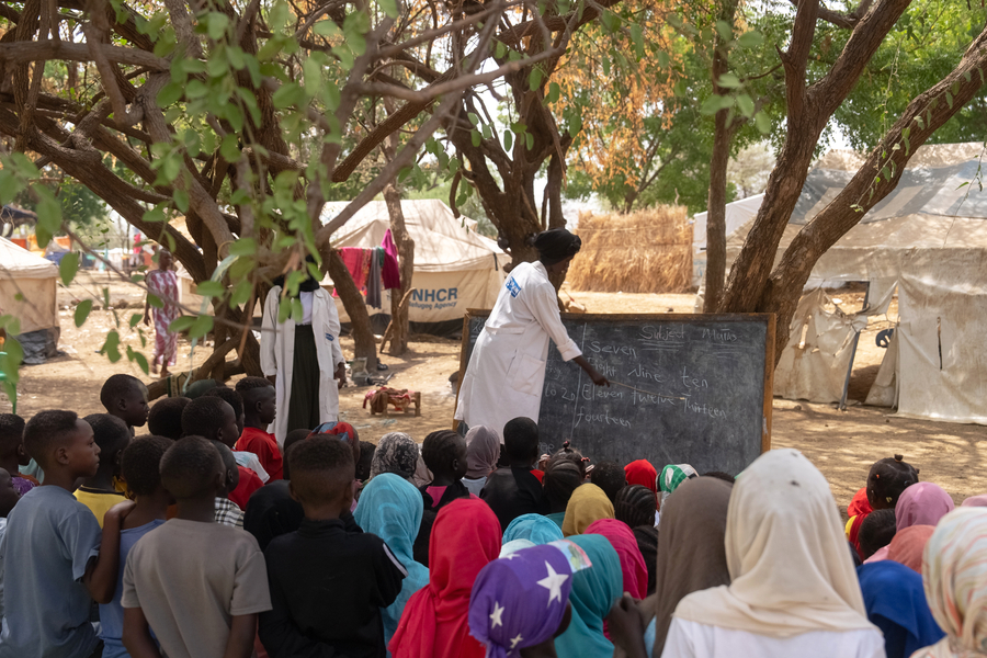 Children are taught math by a teacher pointing at a blackboard under a tree.