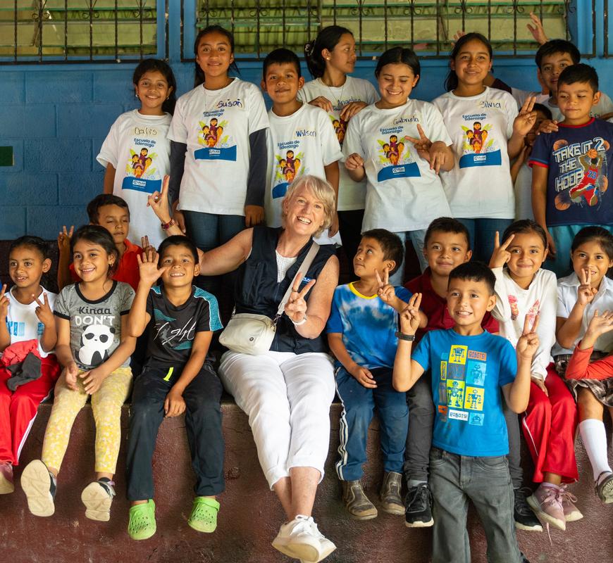 A group of about 20 smiling children holding their hands up and making peace signs, posing with a woman with blonde hair.