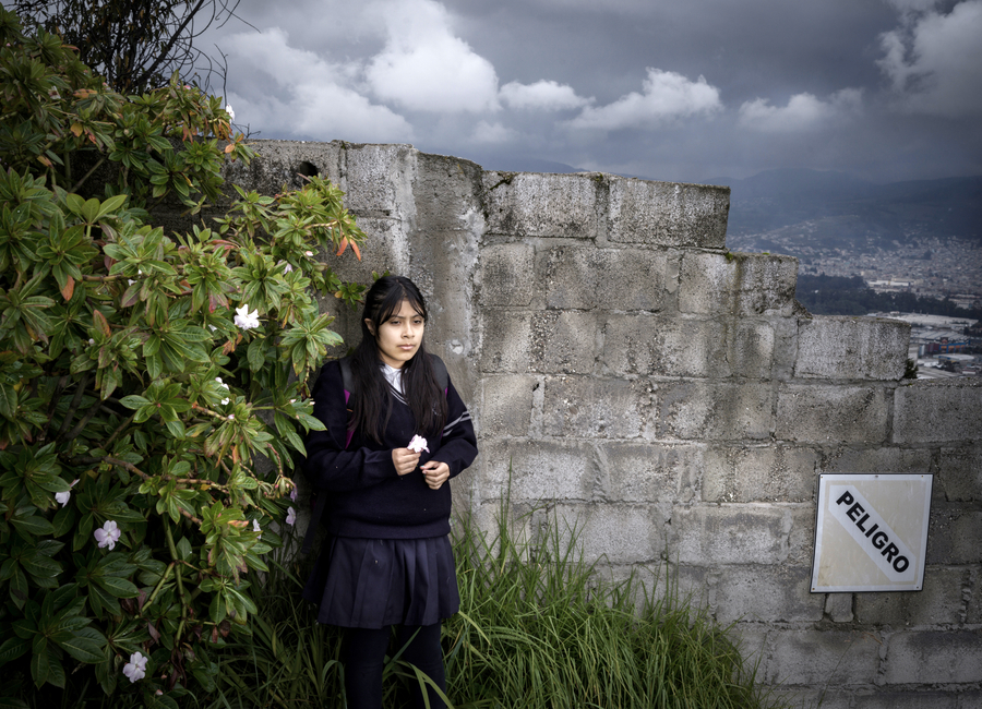 An 11-year-old girl stands by a crumbling wall marked “Peligro,” reflecting concerns about gang violence and insecurity at her school in Ecuador.
