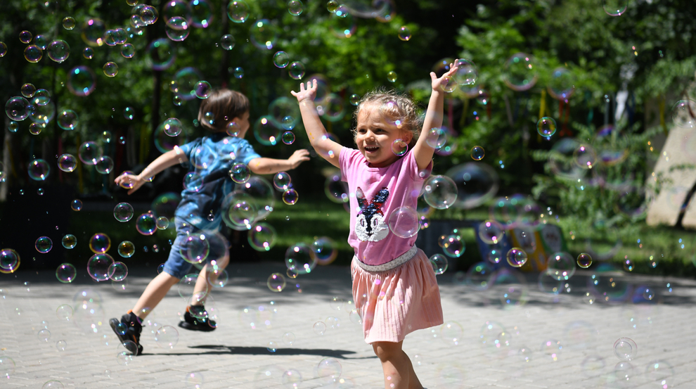 A young girl in a pink shirt and skirt runs through bubbles outside. A boy dressed in blue can be seen in the background.