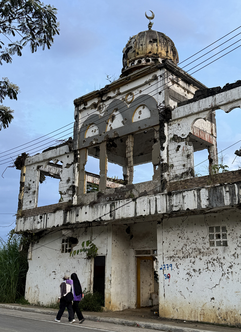 A partially destroyed two-storey building with a weathered dome rises beside a street.