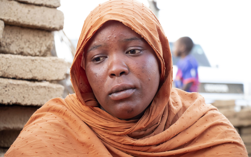 A young woman in an orange headscarf looking downcast at the ground.