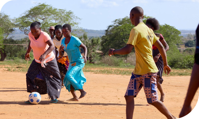 Men and women play soccer together on a dirt field in their bare feet.
