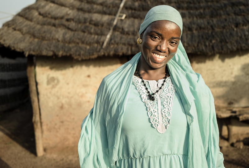 A woman in a green dress and scarf smiles looking directly into the camera. She’s standing in front of her thatched-roof home.