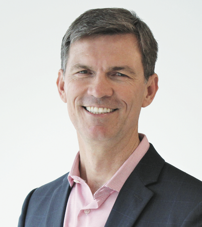 A professional headshot of a man wearing a pink collared shirt with a dark blazer. He is smiling at the camera against a plain light-colored background.