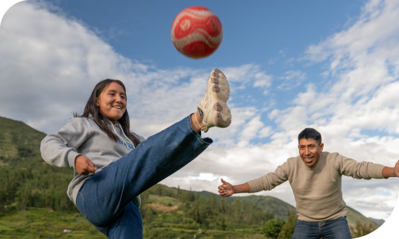 A girl kicks a soccer ball straight up into the air while a man cheers her on.