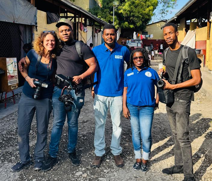 Five adults pose together. A man and a woman on the left and a man on the right carry camera equipment. A man and woman between them wear blue T-shirts with the Plan International logo.