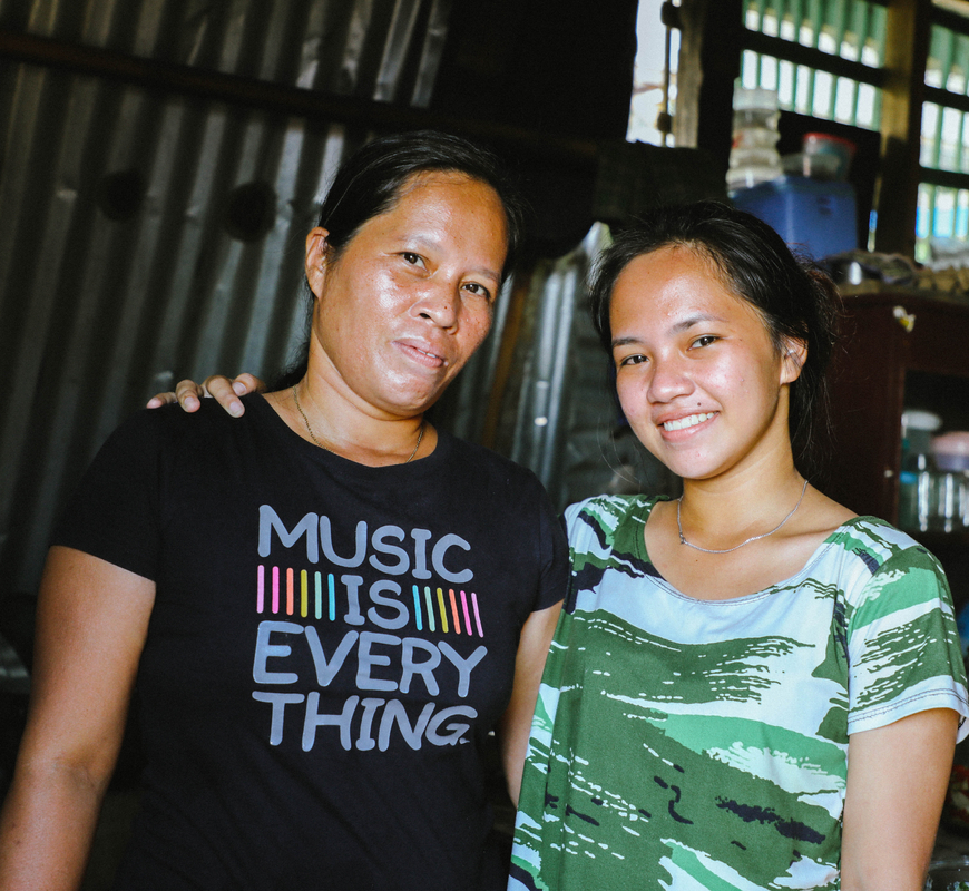 A woman in a black t-shirt and young girl in a green t-shirt stand facing the camera smiling. The young girl has her arm around the woman’s shoulder. 