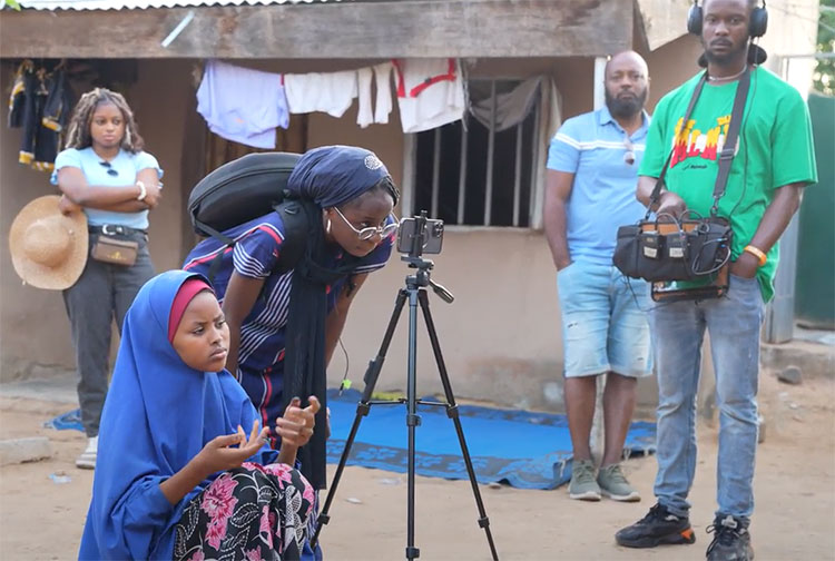 A girl in a blue hijab signs next to a woman filming using a phone on a tripod.
