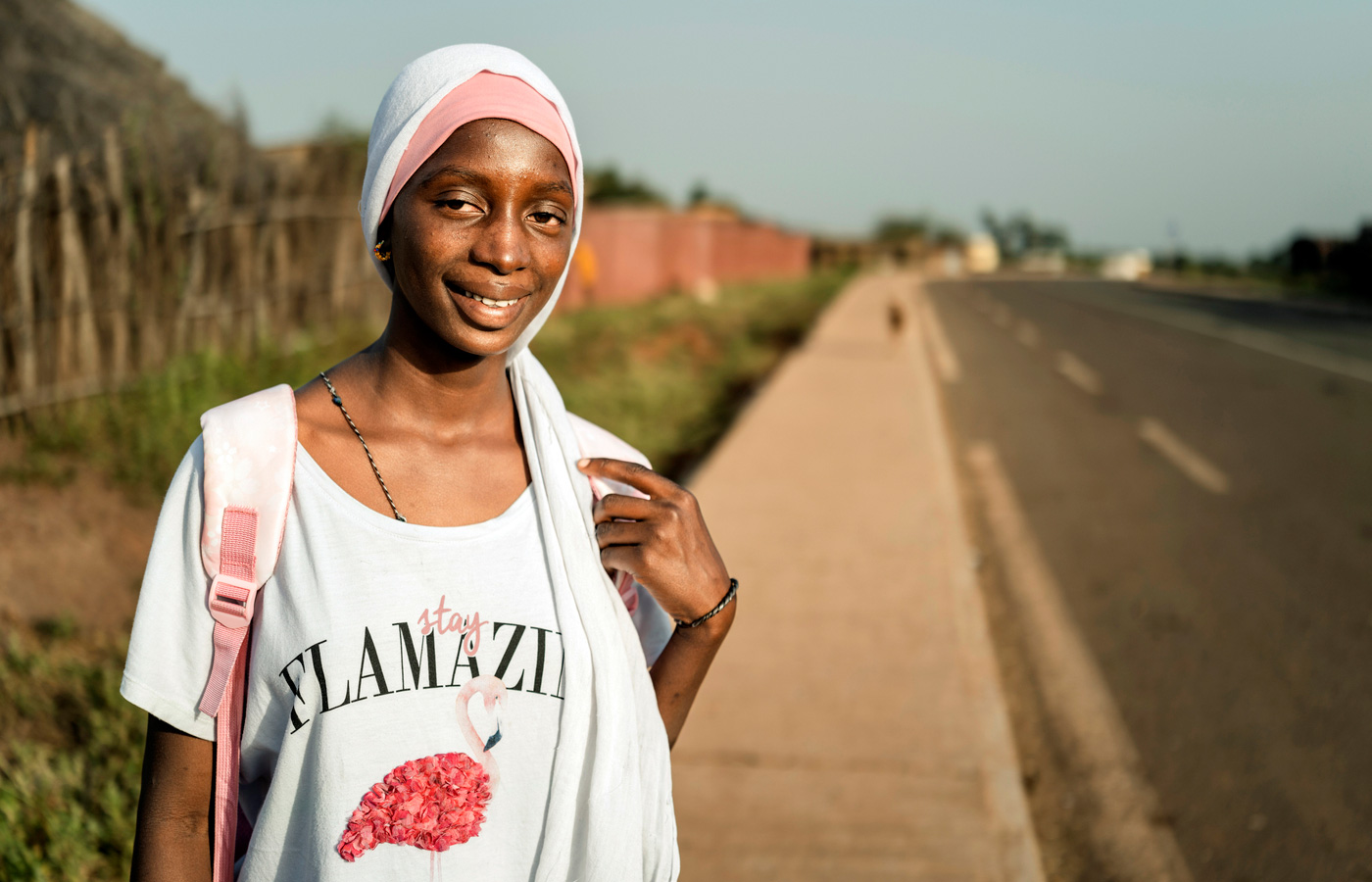 A smiling teenage girl wearing a white and pink head scarf and a white t-shirt stands on the sidewalk beside a road in rural Senegal.