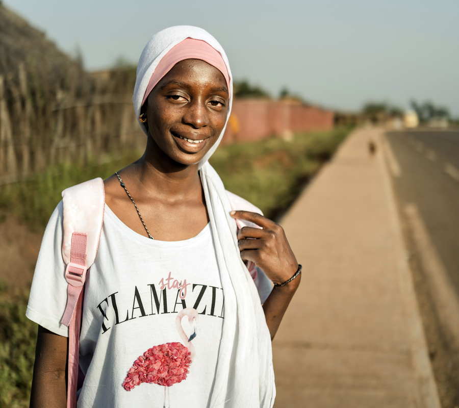 A smiling teenage girl wearing a white and pink head scarf and a white t-shirt stands on the sidewalk beside a road in rural Senegal.