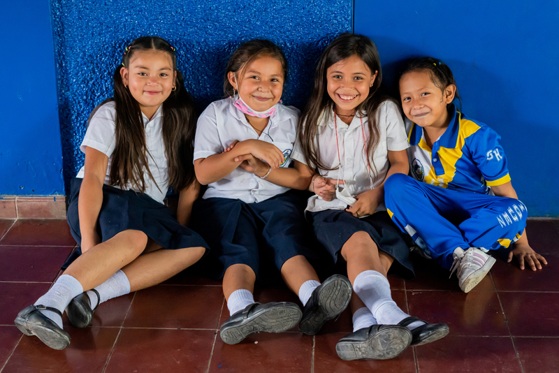 Four girls in school sitting on the ground in front of a blue background, smiling.