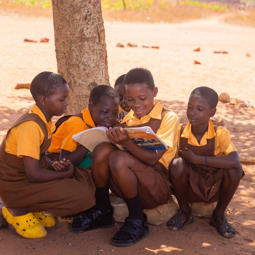 Four children sitting under a tree in a dry, open area, reading a book together. Children are wearing matching yellow shirts and brown uniforms, with one child wearing bright yellow shoes.