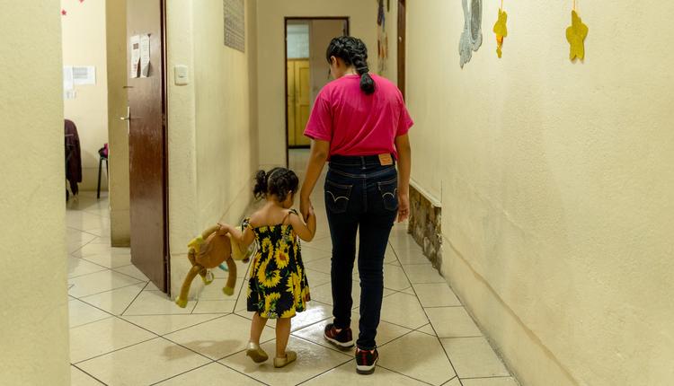 A young woman wearing a pink T-shirt and blue jeans holds a child.