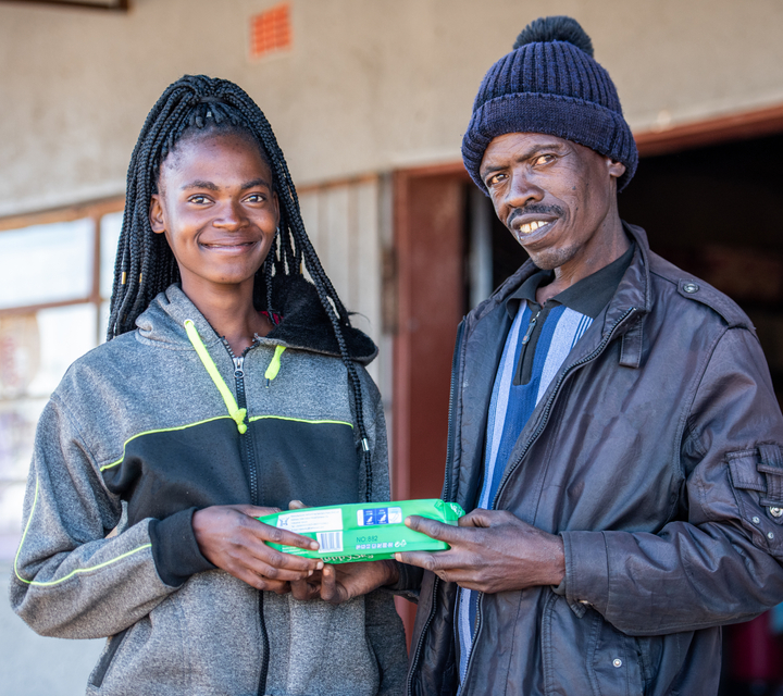 A girl and a man smile and pose for the camera while both holding a box of menstrual pads.