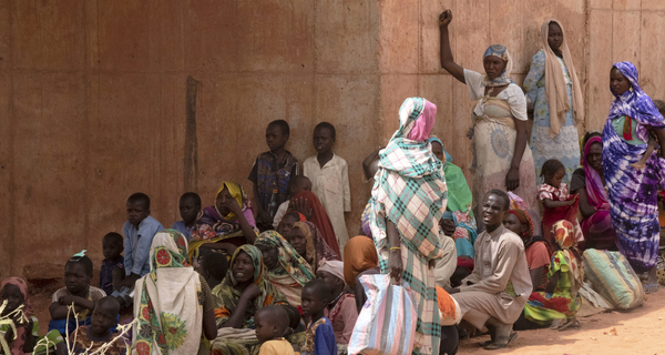 A group of people in Sudan, including adults and children, sit and stand against a large concrete wall, with women wearing colourful patterned clothing and cultural head coverings.