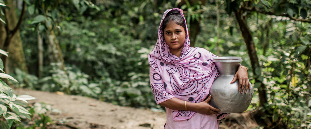 A woman in a pink dress carries a metal pot while standing beside a lush, green path in a forested area.